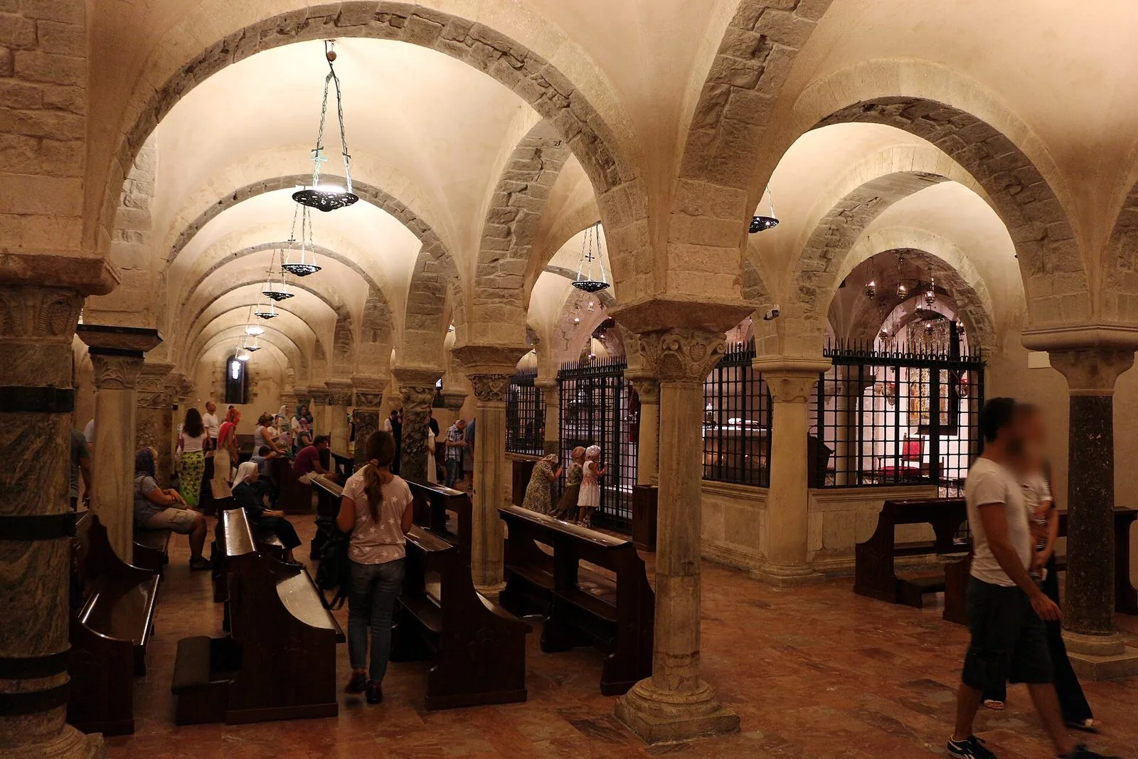 La cripta della Basilica di San Nicola a Bari: volte a crociera su colonne con capitelli scolpiti, panche in legno e fedeli in preghiera davanti alla cancellata della tomba del santo